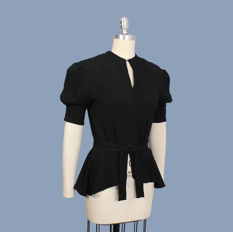 May include: A black, short-sleeved blouse with a keyhole neckline and a fitted waist. The blouse has a peplum hem and a self-tie belt.