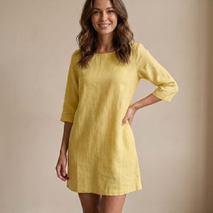 以下が含まれることがあります： 女性が黄色のリネンドレスを着ています。ドレスは丸いネックラインと七分袖です。ドレスは膝丈です。女性は微笑んでいます。