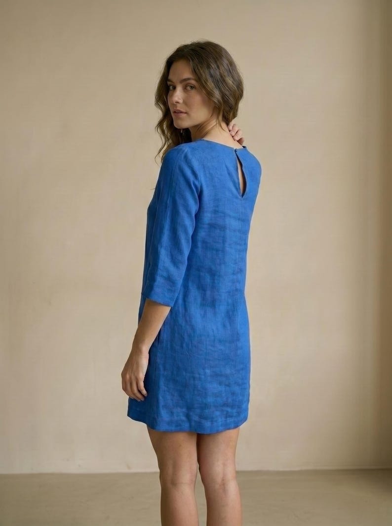 以下が含まれることがあります： 3/4スリーブとキーホールバックが付いた、短い青色のリネンワンピース。ワンピースは無地で、軽量の生地で作られているようです。ワンピースは膝丈です。