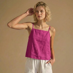 May include: A magenta-colored camisole top with thin straps and a square neckline. The top is paired with white shorts. The outfit is made of a lightweight fabric, perfect for warm weather. The model is fair-skinned with blonde hair.