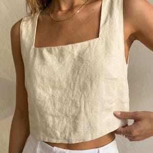 May include: A cream linen crop top with a square neckline. The top is worn with white trousers and a gold necklace. The fabric has a textured appearance. The model is wearing gold rings.