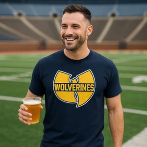 May include: Navy blue t-shirt with a yellow and white graphic that says "WOLVERINES". The person is holding a clear plastic cup with a light-colored beverage. The background is a green field and stadium seating.