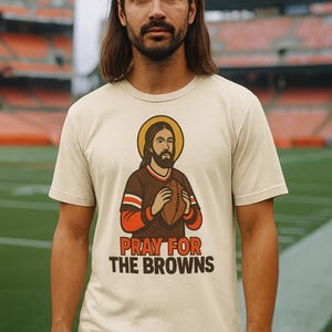 Puede incluir: Camiseta beige con un gráfico de una figura con un halo sosteniendo un balón de fútbol, vistiendo una camiseta marrón y naranja. El texto "PRAY FOR THE BROWNS" está impreso debajo. El fondo muestra un estadio de fútbol.