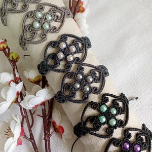May include: Close-up of several macrame bracelets in various colors, including gray, black, and dark gray. Each bracelet features a floral design with pearl-like beads in white, light green, and purple. The bracelets are displayed on a cream-colored surface.