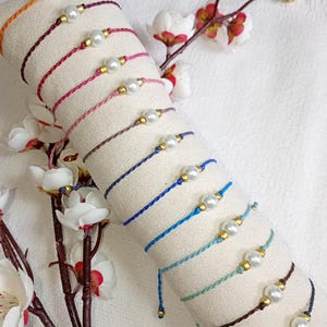 May include: A selection of colourful handmade bracelets, each with a faux pearl and gold-tone beads. The bracelets are displayed on a white fabric roll, with floral decorations. The bracelets are made of braided cord, in a variety of colours.