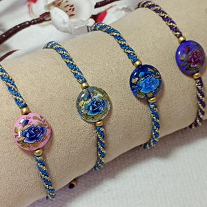 Peut inclure: Quatre bracelets artisanaux avec des motifs floraux. Chaque bracelet présente une perle ronde décorative avec un motif de rose bleue, montée sur un cordon tressé bleu et or. Les perles sont roses, transparentes et bleues, avec des accents dorés.