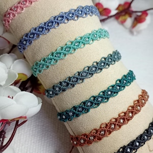 Könnte beinhalten: Eine Auswahl handgefertigter Makramee-Armbänder in verschiedenen Farben, darunter Rosa, Blau, Türkis und Braun. Jedes Armband hat ein gewebtes Design mit kleinen, klaren Perlen. Die Armbänder werden auf beigem Stoff mit floralen Akzenten im Hintergrund präsentiert.