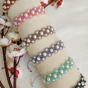 Könnte beinhalten: Eine Auswahl an handgefertigten Makramee-Armbändern in verschiedenen Farben, darunter Braun, Rosa, Lila, Türkis und Schwarz. Jedes Armband hat ein wellenförmiges Design mit weißen Kunstperlen. Die Armbänder werden auf einer zylindrischen, cremefarbenen Oberfläche präsentiert.