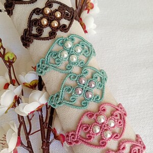 Peut inclure: Gros plan sur plusieurs bracelets en macramé marron, turquoise et rose, chacun orné de perles. Les bracelets sont présentés sur une surface neutre, avec des fleurs blanches en arrière-plan. Les bracelets ont un motif ajouré.