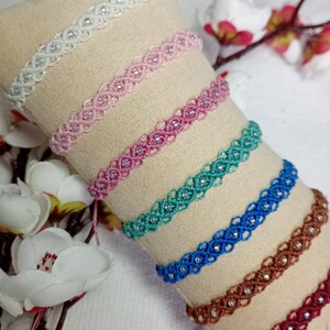 May include: A collection of handmade macrame bracelets in various colors, including white, pink, teal, blue, brown, and burgundy. Each bracelet features a delicate woven design with small, sparkling embellishments. The bracelets are displayed on a beige fabric.