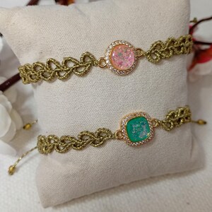 Könnte beinhalten: Zwei goldfarbene Makramee-Armbänder mit verstellbaren Kordeln. Ein Armband hat einen runden, rosa, glitzernden Mittelstein, der von kleinen, klaren Steinen umgeben ist. Das andere hat einen quadratischen, grünen, glitzernden Mittelstein mit einem ähnlichen Rand.