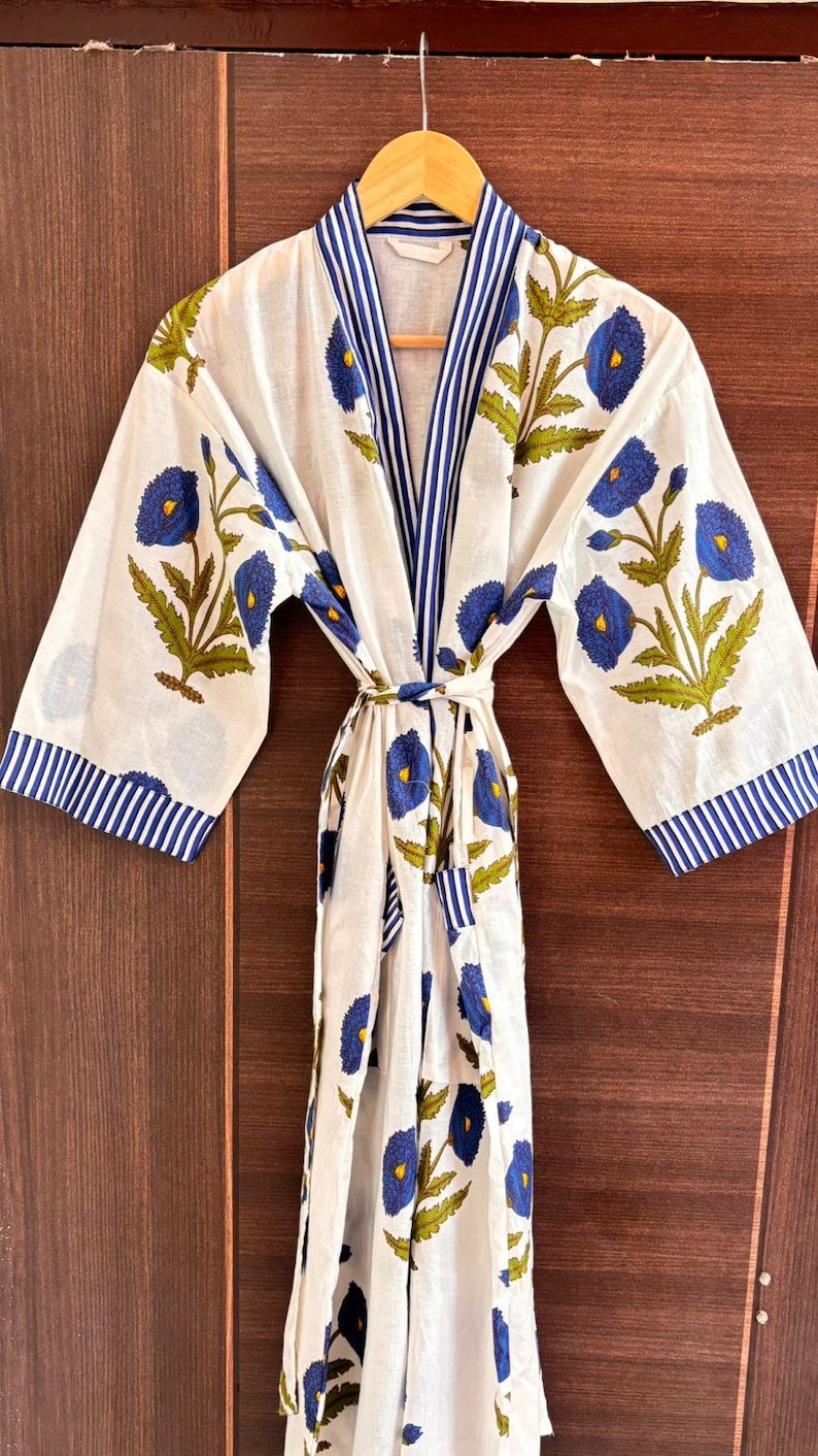 May include: White cotton robe with a blue floral pattern and blue striped trim. The robe features a tie closure and wide sleeves. The floral design includes blue flowers and green leaves. The robe is hanging on a wooden hanger.