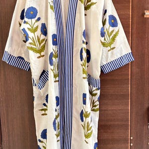 May include: White cotton robe with a blue floral pattern and blue striped trim. The robe features a V-neck, short sleeves, and two pockets. The floral design includes blue flowers and green leaves.