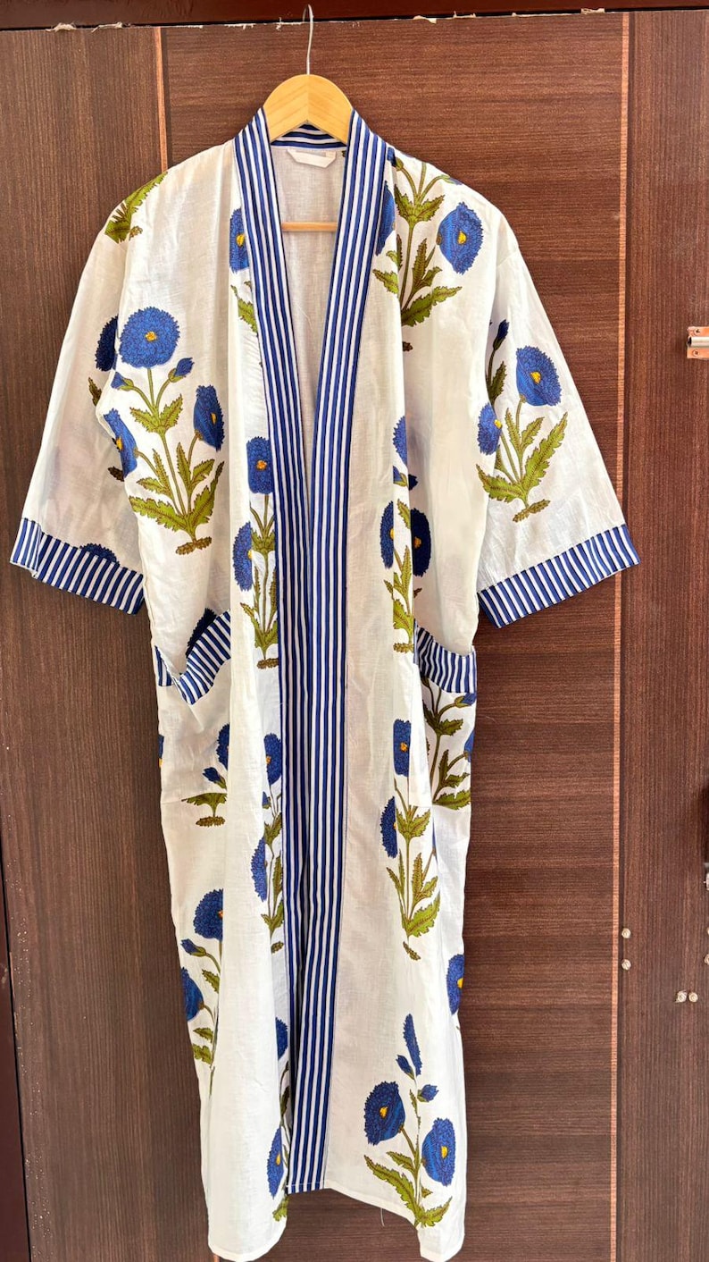 May include: White cotton robe with a blue floral pattern and blue striped trim. The robe features a V-neck, short sleeves, and two pockets. The floral design includes blue flowers and green leaves.