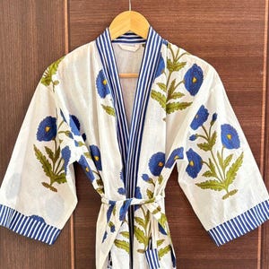 May include: White cotton robe with a blue floral pattern and blue striped trim. The robe features a tie closure and is hanging on a wooden hanger. The floral design includes blue flowers and green leaves.