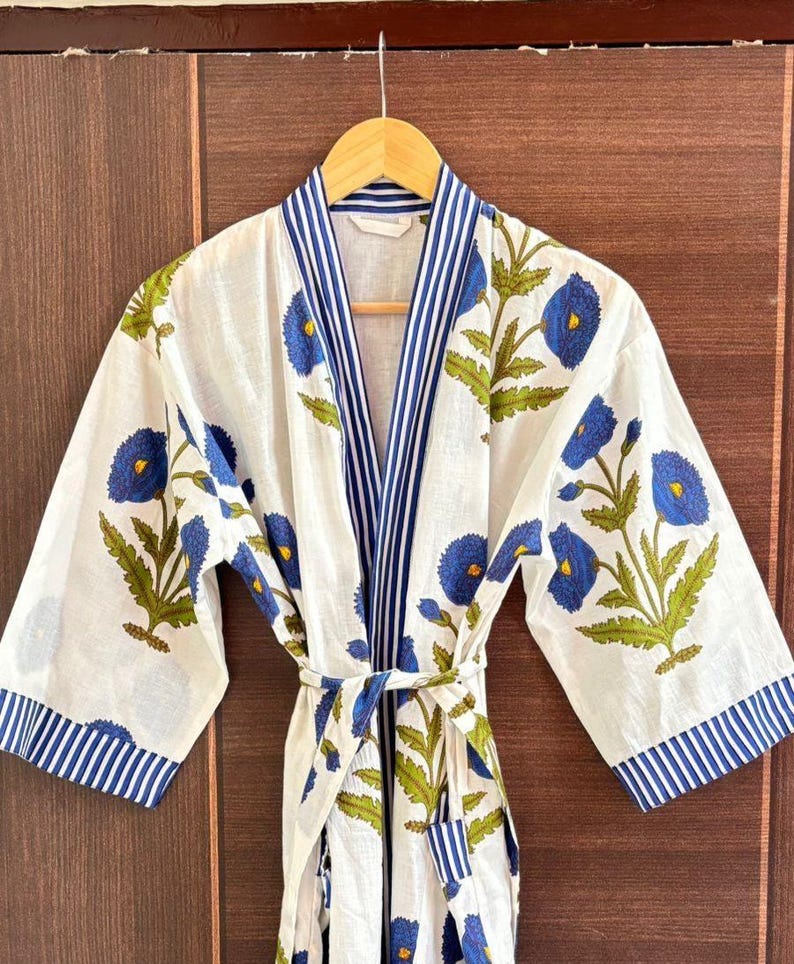 May include: White cotton robe with a blue floral pattern and blue striped trim. The robe features a tie closure and is hanging on a wooden hanger. The floral design includes blue flowers and green leaves.