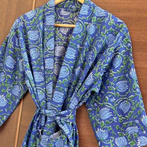 Puede incluir: Bata azul con estampado floral y cinturón a juego, colgada de una percha de madera. La bata presenta un patrón repetido de flores azul claro y hojas verdes sobre un fondo azul más oscuro. La bata está hecha de una tela suave y ligera.
