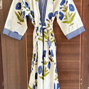 May include: White cotton robe with a blue floral pattern and green leaves. The robe has a striped blue trim on the sleeves and collar, and a matching belt. The robe is hanging on a wooden hanger against a wooden door.