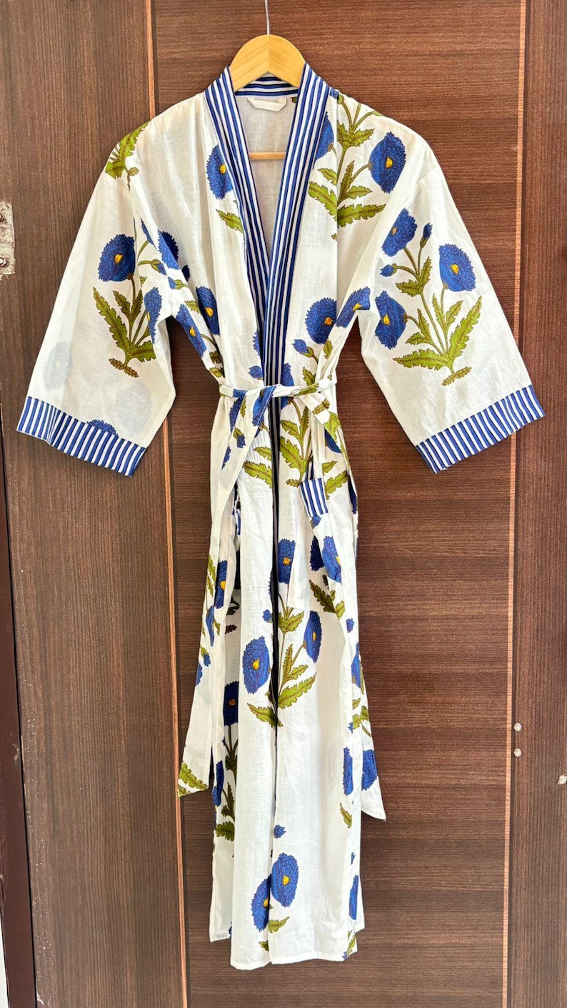 May include: White cotton robe with a blue floral pattern and green leaves. The robe has a striped blue trim on the sleeves and collar, and a matching belt. The robe is hanging on a wooden hanger against a wooden door.