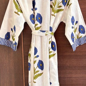 May include: White floral robe with blue and green accents. The robe features a repeating pattern of blue flowers with yellow centers and green leaves. The sleeves and collar have blue and white striped trim. A matching belt cinches the waist.