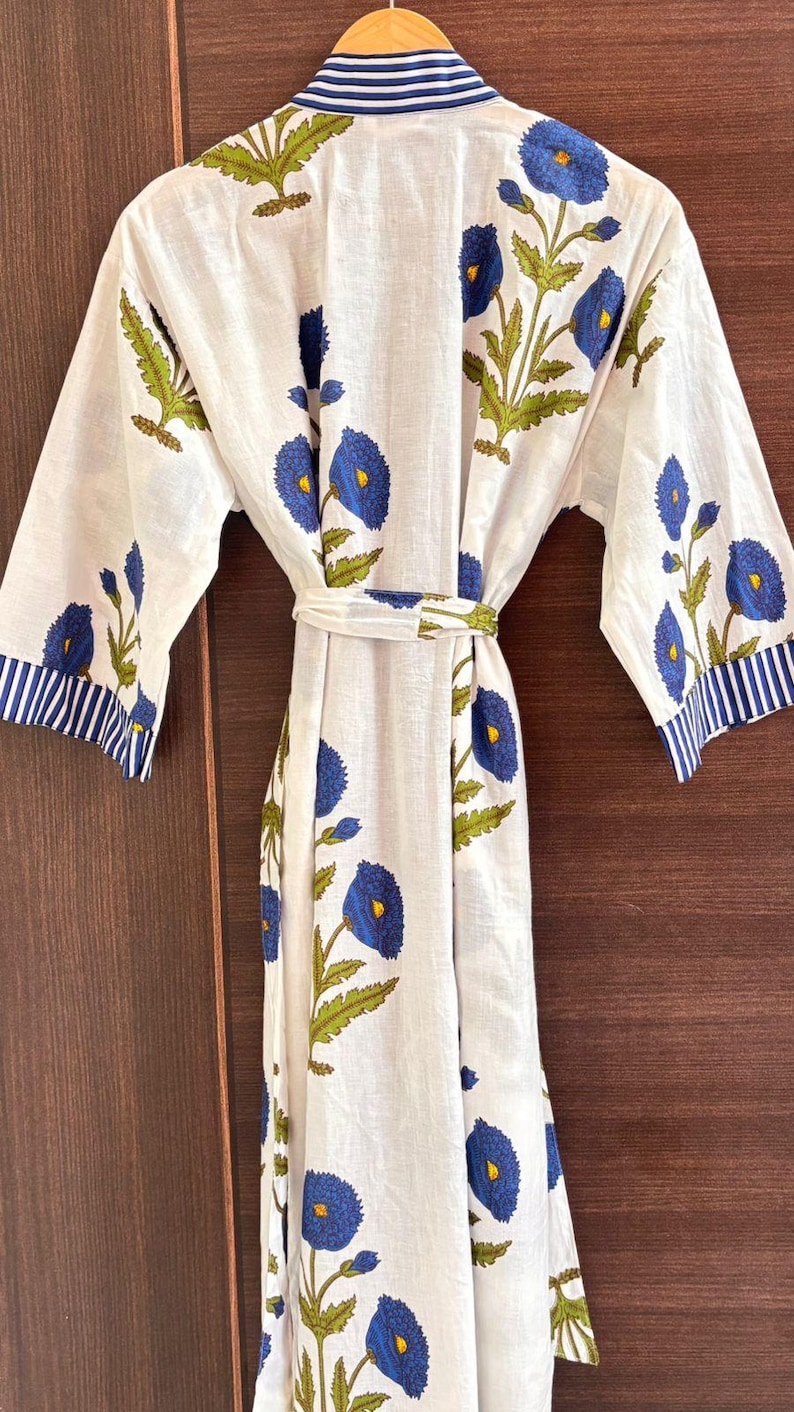 May include: White floral robe with blue and green accents. The robe features a repeating pattern of blue flowers with yellow centers and green leaves. The sleeves and collar have blue and white striped trim. A matching belt cinches the waist.