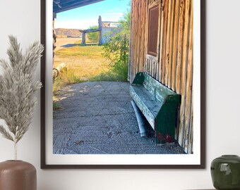 May include: Framed photograph of a rustic wooden building with a green bench. The building has a weathered appearance with vertical wooden planks. The bench is painted green and red. The background shows a field and a blue sky.
