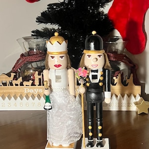 Puede incluir: Dos cascanueces de madera, una novia y un novio, uno al lado del otro. La novia viste un vestido blanco y una corona, sosteniendo una botella de champán. El novio, con uniforme negro, sostiene un ramo y un bastón. Ambos tienen una base con un nombre.