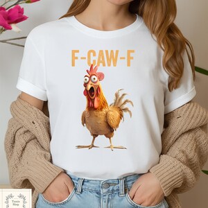 May include: White t-shirt featuring a cartoon chicken with an open beak and the text "F-CAW-F" in orange. The chicken is brown, orange, and red with wide eyes. The shirt has rolled-up sleeves.
