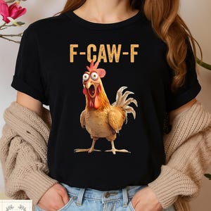 May include: Black t-shirt featuring a cartoon chicken with wide eyes and an open beak. The text "F-CAW-F" is printed above the chicken in a gold color. The shirt is a crew neck style.