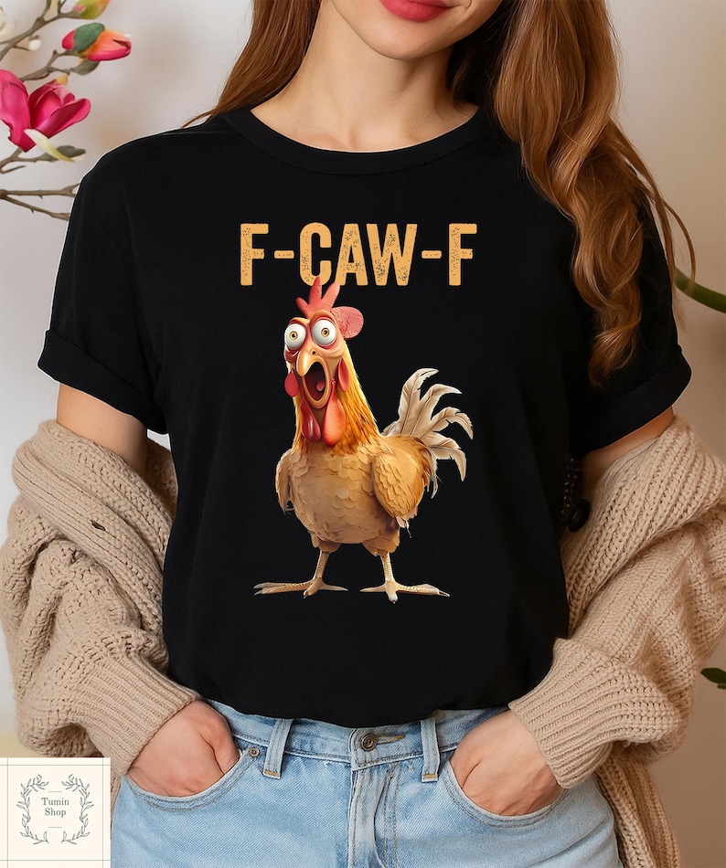 May include: Black t-shirt featuring a cartoon chicken with wide eyes and an open beak. The text "F-CAW-F" is printed above the chicken in a gold color. The shirt is a crew neck style.