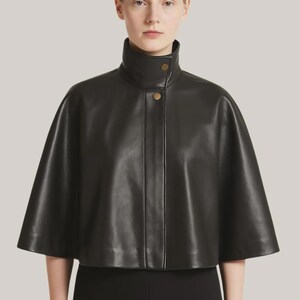 May include: A black leather cape with a high, rounded collar and two gold-colored buttons. The cape has a short, flared silhouette and is made of a smooth, shiny material. The garment is part of a fashion collection.