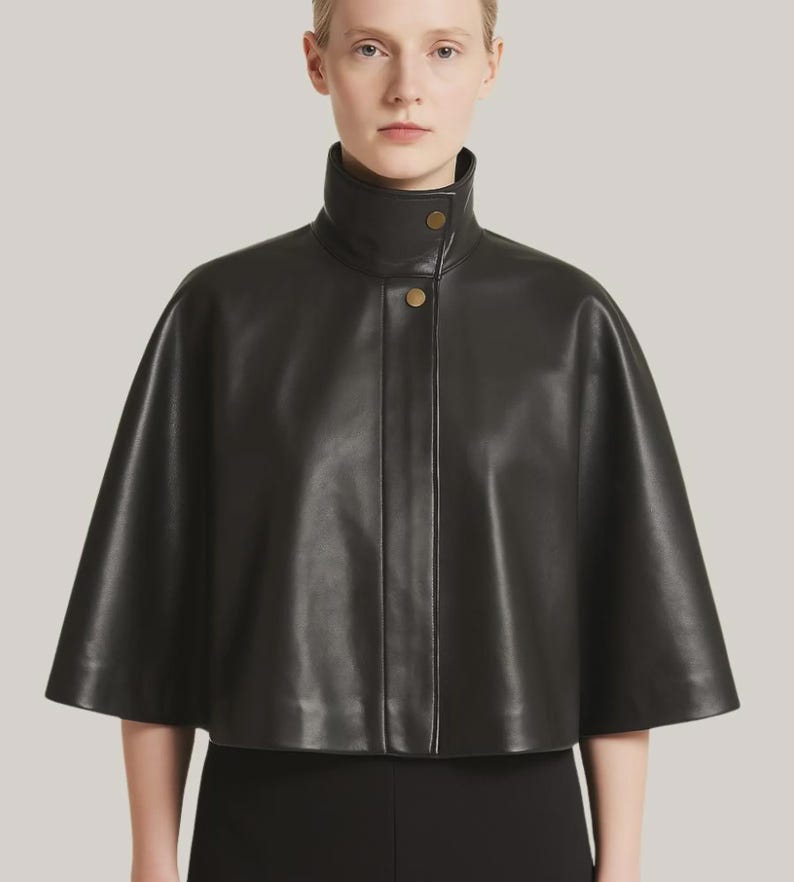 May include: A black leather cape with a high, rounded collar and two gold-colored buttons. The cape has a short, flared silhouette and is made of a smooth, shiny material. The garment is part of a fashion collection.