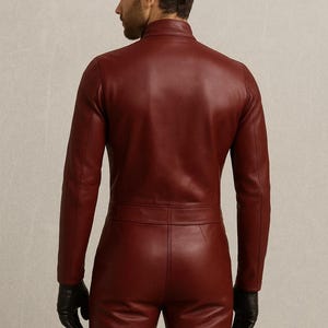 May include: A burgundy leather jumpsuit with a high collar and long sleeves. The one-piece garment features a fitted silhouette, a belt detail at the waist, and matching burgundy leather pants. The model is wearing black gloves.
