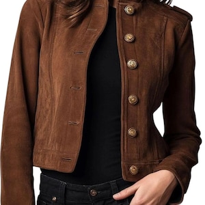 May include: A brown suede jacket with a high collar and gold buttons. The jacket has a tailored fit and is worn over a black top and black jeans. The jacket has shoulder epaulets and a classic design.