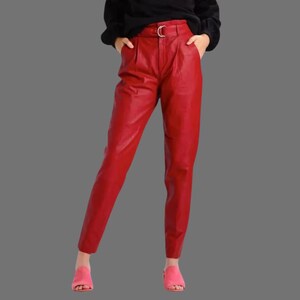 May include: Red leather pants with a high waist and a gold-colored belt buckle. The pants have a tailored fit and are slightly tapered at the ankle. The model is wearing pink sandals.