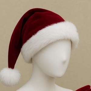 Puede incluir: Un gorro de Papá Noel clásico de terciopelo rojo oscuro con un ribete de piel sintética blanca y un pompón a juego. El gorro se exhibe en una cabeza de maniquí, destacando su diseño tradicional y su atractivo festivo. Perfecto para celebraciones navideñas.