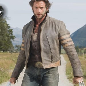 May include: A gray leather jacket with tan stripes on the sleeves and a zipper closure. The jacket is worn over a brown shirt and blue jeans. The person is holding metal claws. The background features a dirt path and mountains.