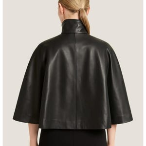 May include: A black leather cape with a high, rounded collar. The cape has short sleeves and a smooth, shiny texture. The garment is seen from the back, showcasing its tailored design and elegant silhouette. The cape is a stylish outerwear piece.