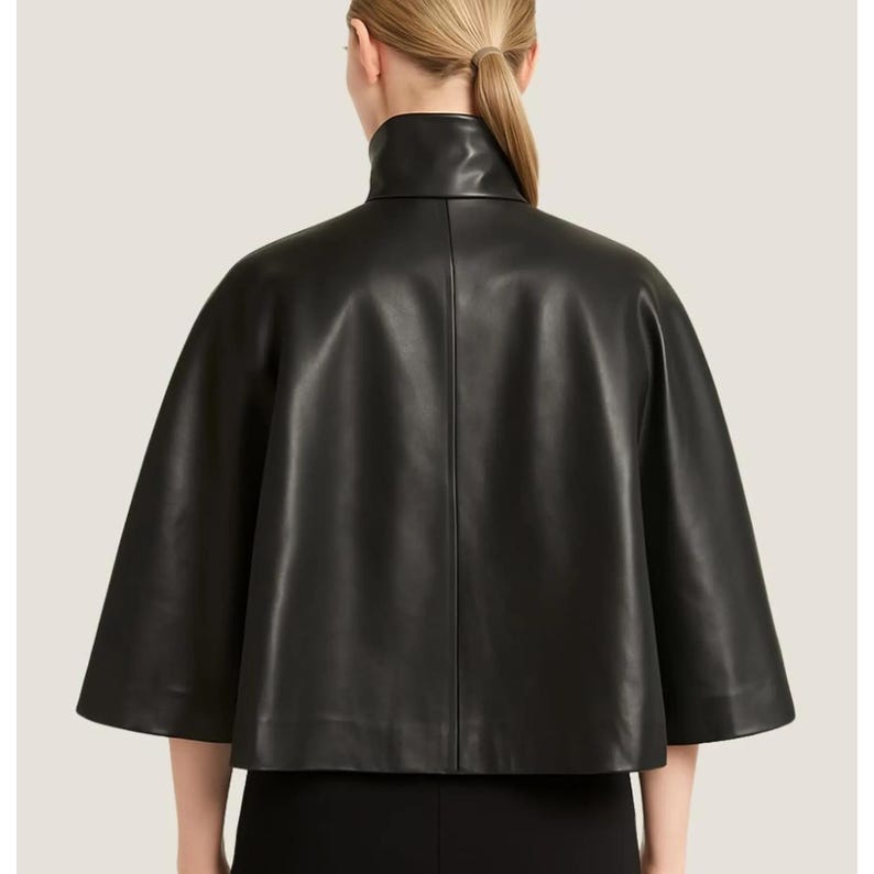 May include: A black leather cape with a high, rounded collar. The cape has short sleeves and a smooth, shiny texture. The garment is seen from the back, showcasing its tailored design and elegant silhouette. The cape is a stylish outerwear piece.
