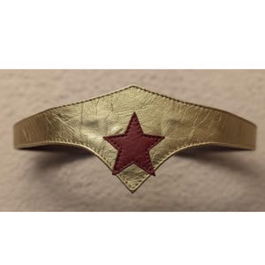 May include: Gold-colored headband with a red star emblem. The headband has a metallic finish and a stitched design. The star is centered on the front of the headband, which has a pointed bottom edge.