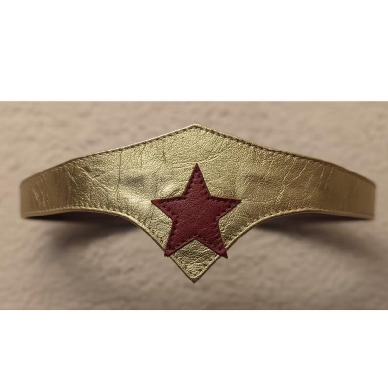 May include: Gold-colored headband with a red star emblem. The headband has a metallic finish and a stitched design. The star is centered on the front of the headband, which has a pointed bottom edge.