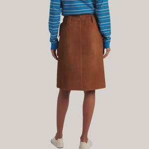 May include: A brown suede skirt with a belt and a knee-length hemline. The skirt is paired with a blue and white striped long-sleeve shirt and white sneakers. The skirt is a classic, versatile piece.