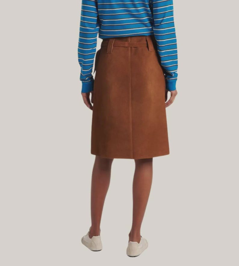 May include: A brown suede skirt with a belt and a knee-length hemline. The skirt is paired with a blue and white striped long-sleeve shirt and white sneakers. The skirt is a classic, versatile piece.