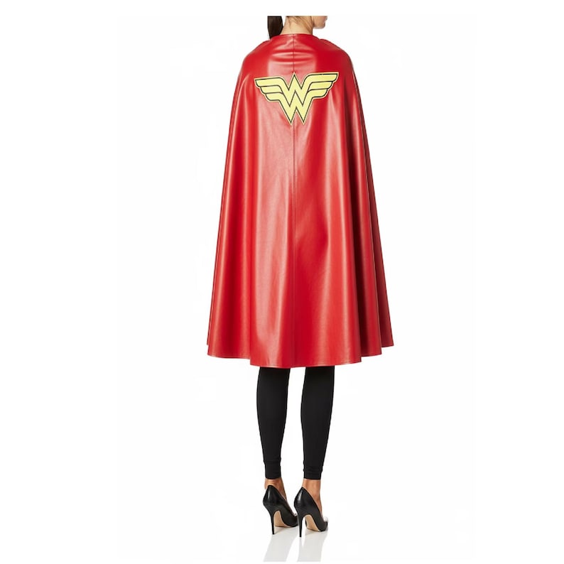 May include: A red cape with a yellow emblem on the back. The cape is made of a shiny material and is knee-length. The emblem is a stylized "W" with wings. The person is wearing black leggings and black high heels.