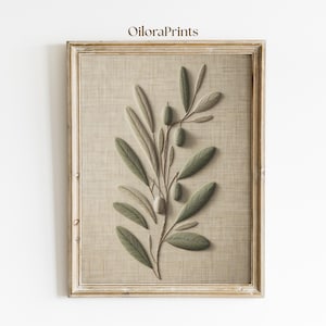 May include: A framed print of an olive branch with green leaves and olives, set against a beige linen-textured backdrop. The artwork is enclosed in a light brown wooden frame. The text "OiloraPrints" is in the top left corner of the image.