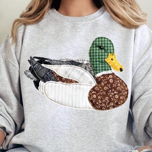May include: A light grey sweatshirt with a patchwork duck design. The duck has a green checked head, a yellow beak with black polka dots, and wings with various patterned fabrics. The duck's body is brown with a floral pattern.