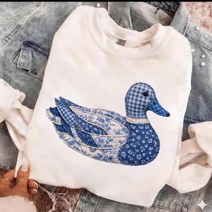 Könnte beinhalten: Weißes Sweatshirt mit einem Enten-Applikation. Das Enten-Design verwendet blaue Blumen- und Vichy-Muster, was einen Patchwork-Effekt erzeugt. Das Sweatshirt wird auf einer Jeansjacke gezeigt. Maße nicht verfügbar.