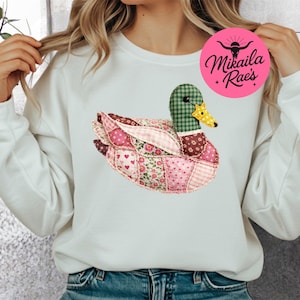 Può includere: Felpa bianca con un design a patchwork di anatra. L'anatra ha una testa a scacchi verdi, un becco giallo e un corpo in tessuto rosa a motivi floreali e a cuore. La felpa ha anche un logo circolare rosa con la scritta "Mikaila Rae's".
