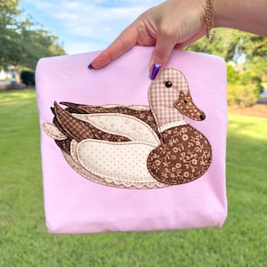 May include: A pale pink sweatshirt with an appliqued duck design. The duck is made from patchwork fabrics with brown floral, checked, and dotted patterns. The duck's beak is brown, and the eye is black. The sweatshirt is held up by a hand.