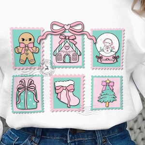 May include: White sweatshirt featuring six Christmas-themed postage stamp designs. The stamps depict a gingerbread man, a house, a snow globe, a wrapped gift, a stocking, and a Christmas tree, all in pastel colors. A pink bow connects the top three stamps.
